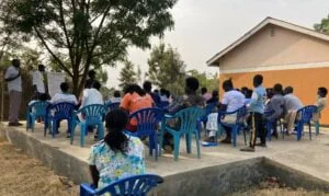 Kuva on Ugandan talouslukutaidon kurssin opetustilateesta. Joukko ihmisiä istuu sinisillä tuoleilla ulkotilassa ja katsovat opetustaululle