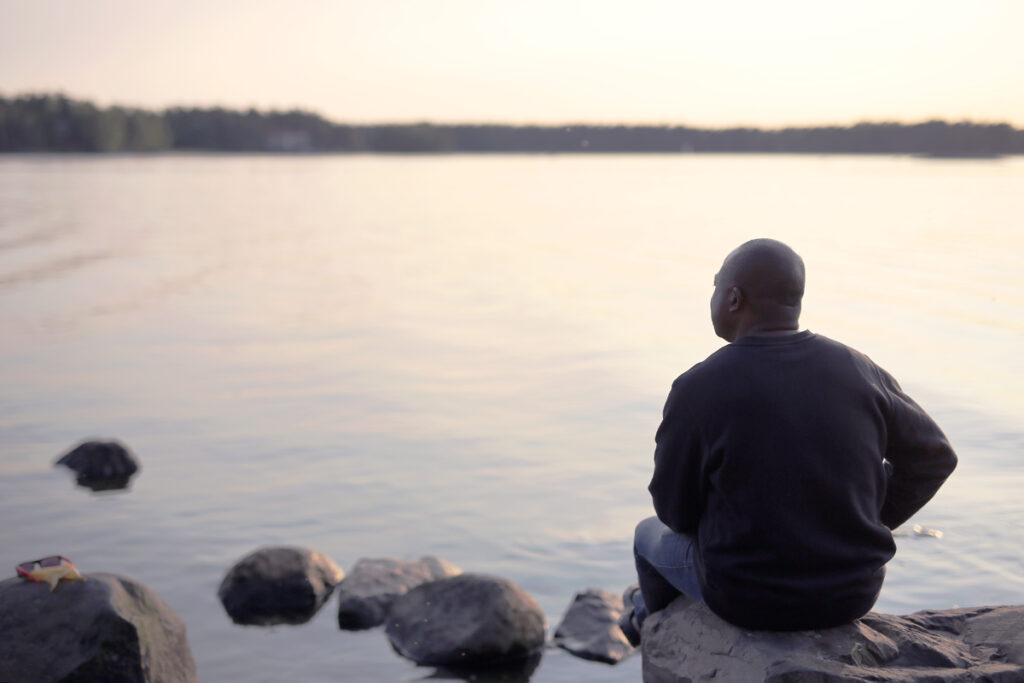 Mies istuu kivellä järven rannalla selkä kameraan päin.