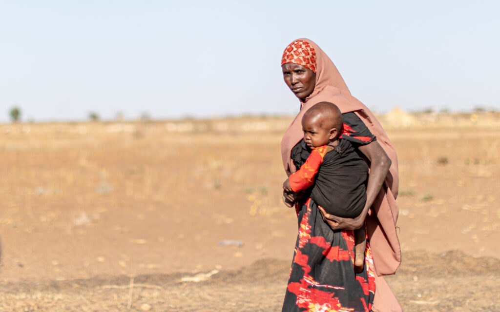 Etiopialainen huvipäinen nainen kantaa sylissään vauvaa. Taustalla näkyy kuivaa aavikkoa ja vaaleansininen taivas.