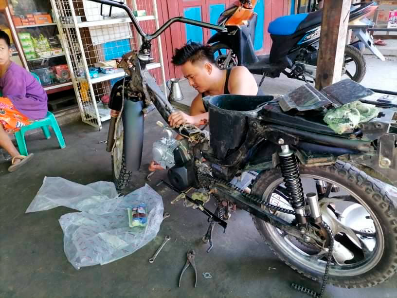 Hihattomaan paitaan pukeutunut mustahiuksinen mies korjaa moottoripyörää. Etualalla näkyy työkaluja ja sivulla nuori henkilö, joka istuu pienellä jakkaralla.