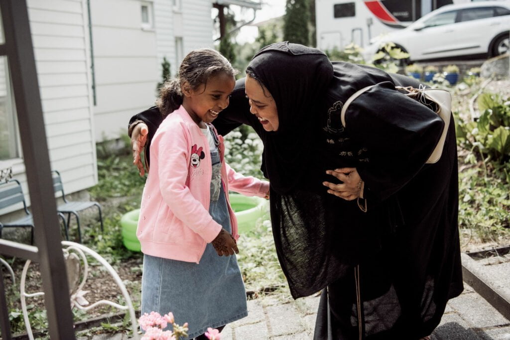 Mustaan asuun pukeutunut nainen kumartuu halaamaan pientä tyttöä, jolla on yllään vaaleanpunainen neuletakki. Molemmat nauravat. Taustalla näkyy kesäinen pihaympäristö.