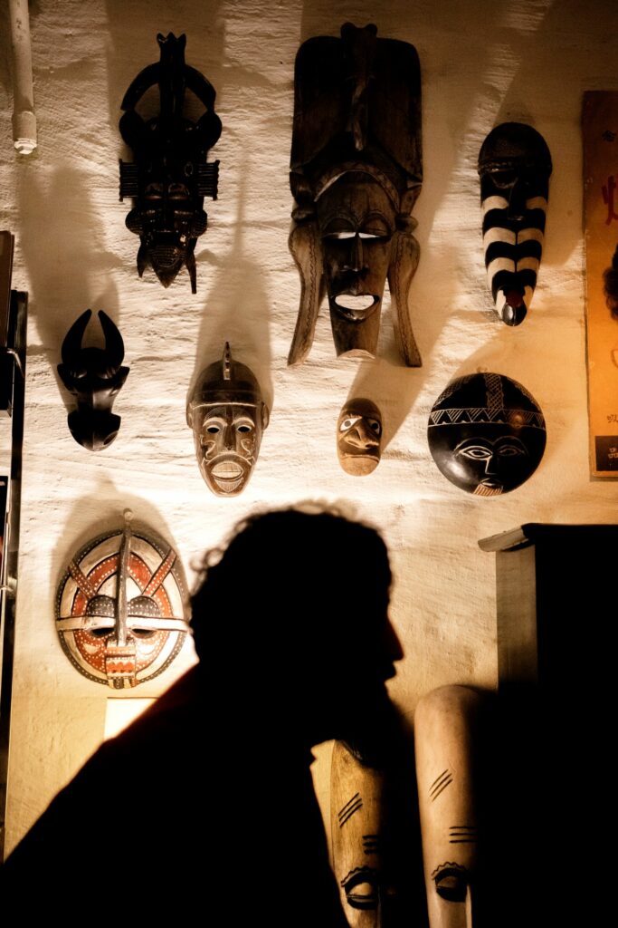 Kuvassa näkyy hämärässä valaistuksessa miehen sivuprofiili. Taustalla seinä, johon on kiinnitetty kahdeksan eri kokoista afrikkalaista maskia.