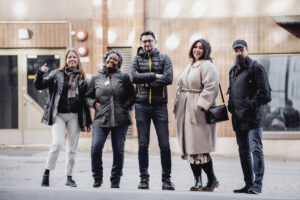 Kuvassa viisi iloista ihmistä seisoo kadulla ulkona.