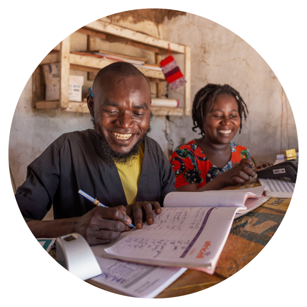 Kuvassa on hymyilevä mies ja nainen Ugandassa. Henkilöt täyttävät laskimen avulla pöydällä olevaa tilikirjaa