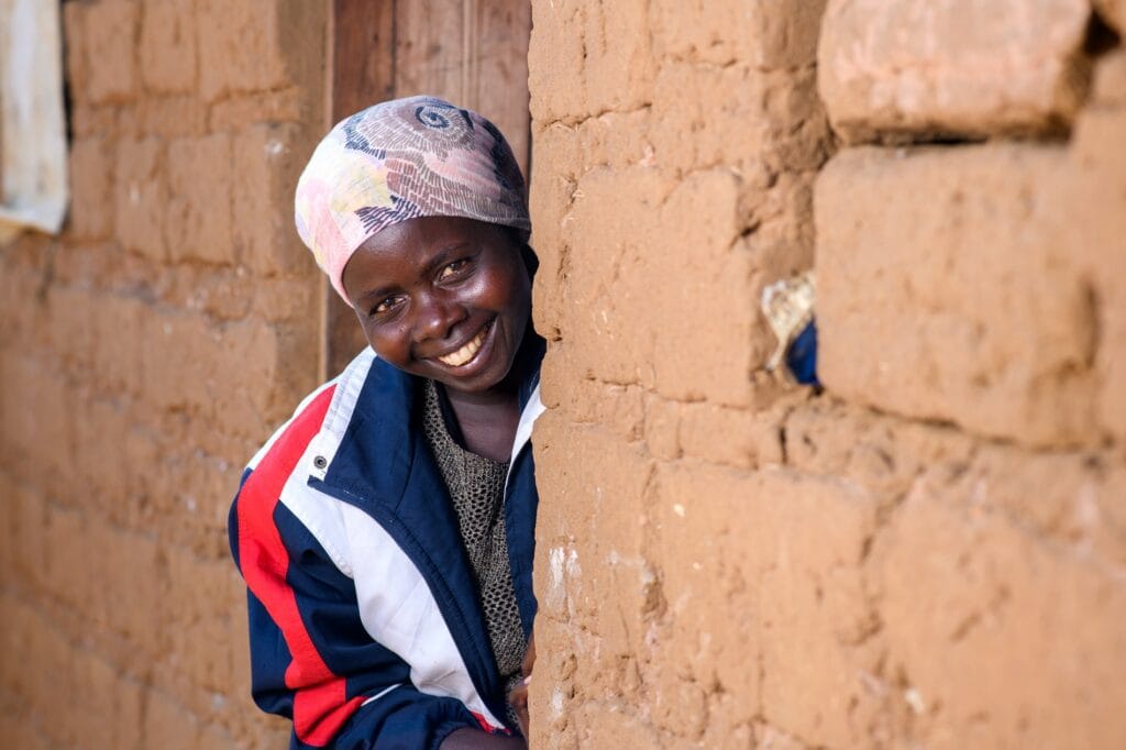 Ugandalainen nainen nimeltä Everine kurkistaa talon nurkan takaa ja hymyilee.