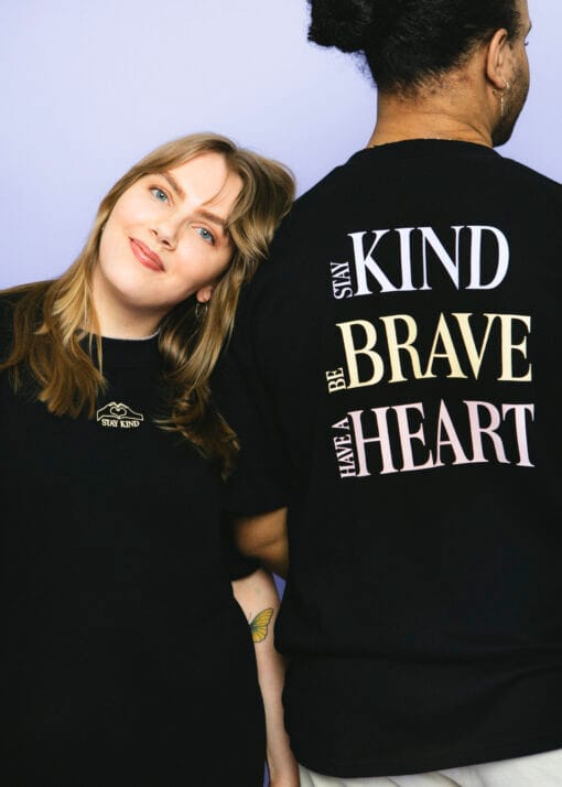 Nainen katsoo kameraan ja hymyilee. Naisen vieressä on mies, joka on selin kameraan. Molempien ihmisten päällä on Pakolaisavun Stay Kind -paita, jonka etupuolella lukee pienellä Stay Kind ja takapuolella isommalla Stay Kind, Be Brave, Have a Heart.