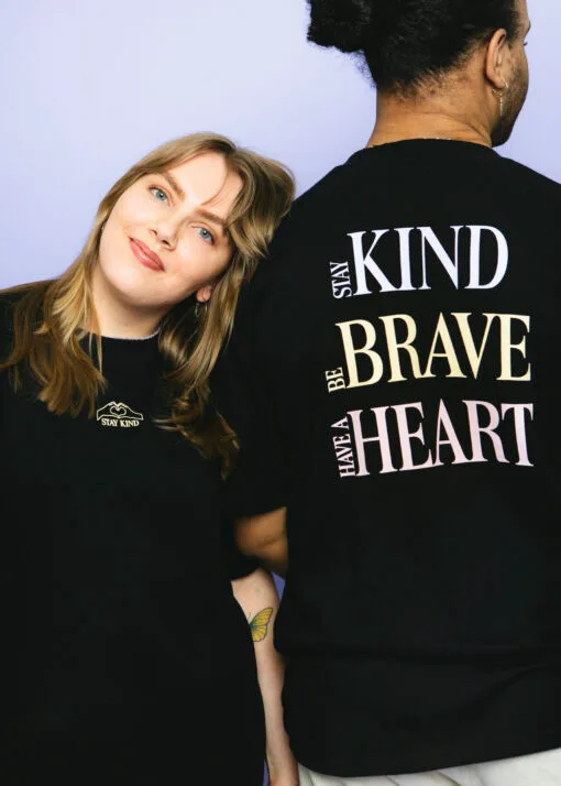Nainen katsoo kameraan ja hymyilee. Naisen vieressä on mies, joka on selin kameraan. Molempien ihmisten päällä on Pakolaisavun Stay Kind -paita, jonka etupuolella lukee pienellä Stay Kind ja takapuolella isommalla Stay Kind, Be Brave, Have a Heart.