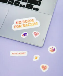 Kuvassa näkyy violettia taustaa vasten kannettavan tietokoneen näppäimistö, johon on liimattu eri värisiä ja eri kokoisia kuva- ja tekstitarroja. Suurimmassa tarrassa lukee No room for racism