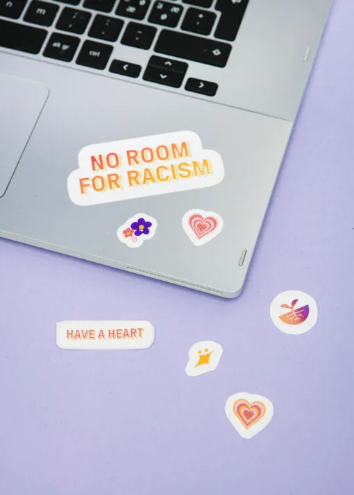Kuvassa näkyy violettia taustaa vasten kannettavan tietokoneen näppäimistö, johon on liimattu eri värisiä ja eri kokoisia kuva- ja tekstitarroja. Suurimmassa tarrassa lukee No room for racism